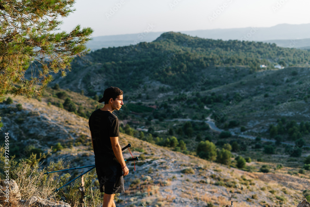 Side view of active young male with hiking poles standing on mountain slope and enjoying landscape while taking break during trekking workout Side view of active young male with hiking poles standing on mountain slope and enjoying landscape while taking break during trekking workout