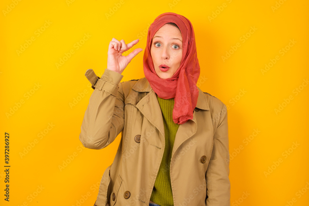 Shocked Young caucasian Muslim woman wearing hijab standing against yellow wall shows something little with hands, demonstrates size, opens mouth from surprise. Measurement concept. Shocked Young caucasian Muslim woman wearing hijab standing against yellow wall shows something little with hands, demonstrates size, opens mouth from surprise. Measurement concept.