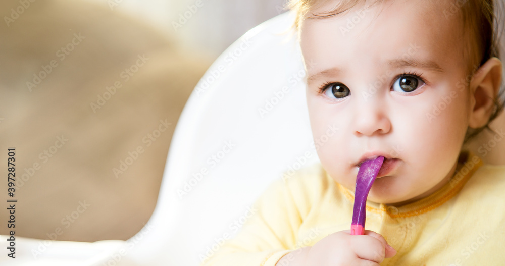 Little baby sitting in high chair and eating food. baby's first feeding. 6 month old toddler holding spoon in mouth Little baby sitting in high chair and eating food. baby's first feeding. 6 month old toddler holding spoon in mouth