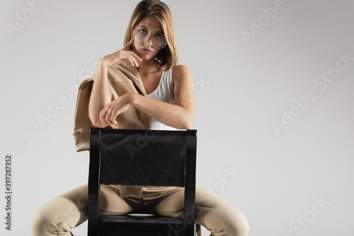 A cute girl in a white T-shirt, pants, glasses, sits on a chair in the studio without emotion, thinks