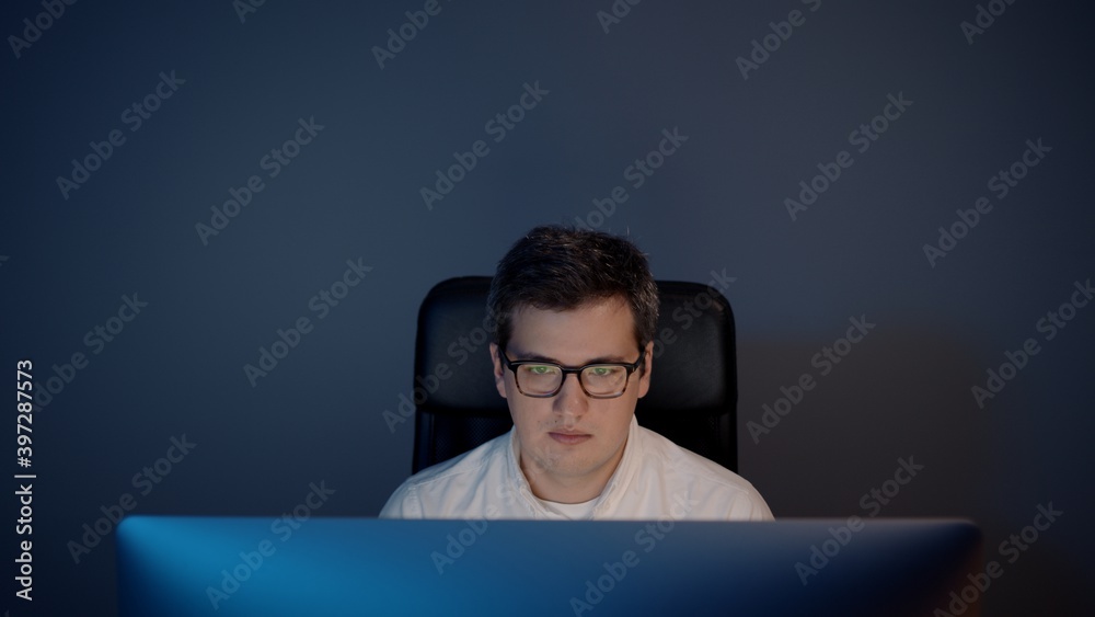Man in eyeglasses in front of the computer in home office on background of grey wall. Pan shot of young man sitting at home working alone at night Man in eyeglasses in front of the computer in home office on background of grey wall. Pan shot of young man sitting at home working alone at night