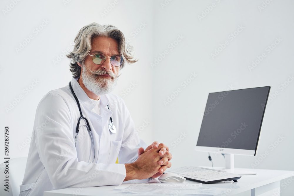 Portrait of senior male doctor with grey hair and beard in white coat is indoors in clinic Portrait of senior male doctor with grey hair and beard in white coat is indoors in clinic