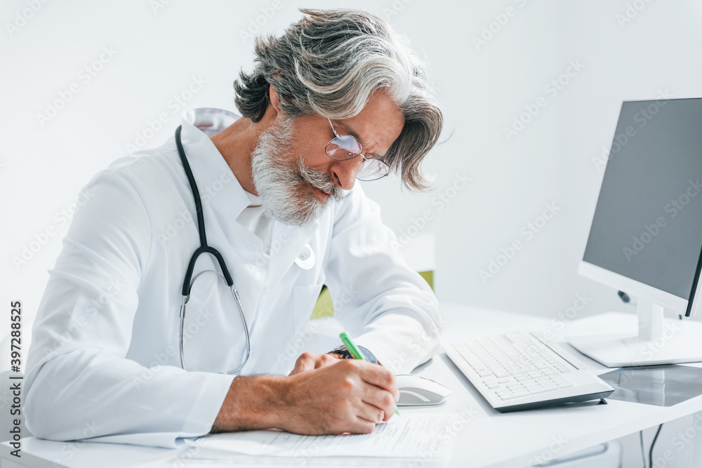 Writes by computer table. Senior male doctor with grey hair and beard in white coat is indoors in clinic Writes by computer table. Senior male doctor with grey hair and beard in white coat is indoors in clinic
