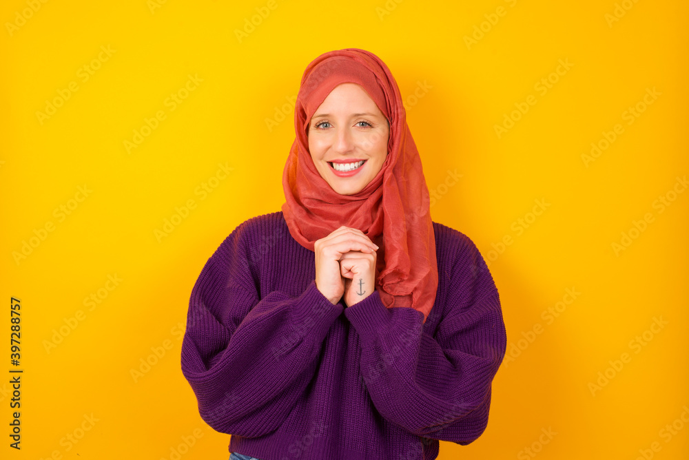 Dreamy charming Young caucasian Muslim woman wearing hijab standing against yellow wall with pleasant expression, keeps hands crossed near face, excited about something pleasant. Dreamy charming Young caucasian Muslim woman wearing hijab standing against yellow wall with pleasant expression, keeps hands crossed near face, excited about something pleasant.