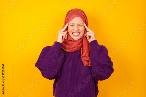 Young caucasian Muslim woman wearing hijab standing against yellow wall concentrating hard on an idea with a serious look, thinking with both index fingers pointing to forehead. © Jihan