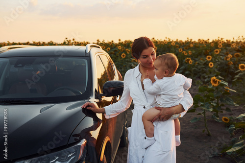 Near modern black car. Young mother with her little son is outdoors in the agricultural field. Beautiful sunshine
