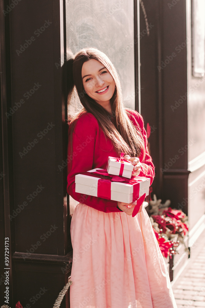 Young cheerful beautiful woman in pink skirt, red sweater hold gift boxes in hand. smile outdoor. Happy attractive cute girl with Valentines day present on romantic city street. Celebrate love holiday Young cheerful beautiful woman in pink skirt, red sweater hold gift boxes in hand. smile outdoor. Happy attractive cute girl with Valentines day present on romantic city street. Celebrate love holiday