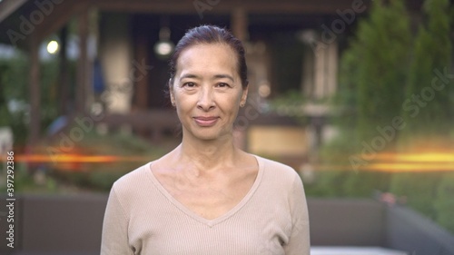 Asian woman on background of private house, looking at the camera, blurred background, lens flare. Adult woman owner of a modern private country house backyard