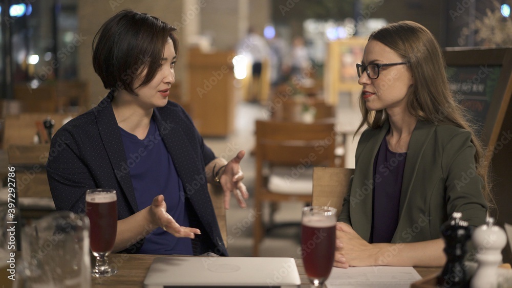 Two women friends sitting at the bar, blond woman in eyeglasses sitting on background of empty chairs. Two women discussing in an open space bar