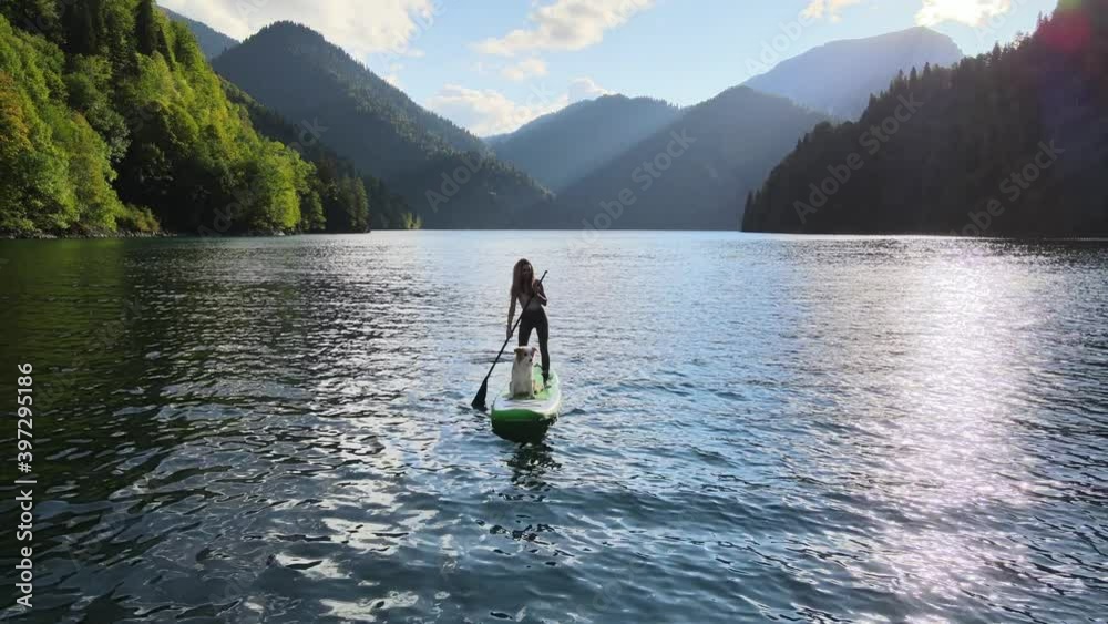 Aerial drone view of pretty fit woman paddle on sup board at mountain lake during sunset with her dog. Concept of active tourism. Female silhouette doing water sport during summer holidays. Aerial drone view of pretty fit woman paddle on sup board at mountain lake during sunset with her dog. Concept of active tourism. Female silhouette doing water sport during summer holidays.