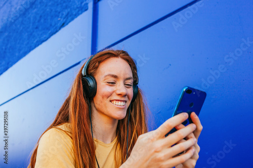 Redhead woman wearing headphones using mobile phone while sitting against blue wall
