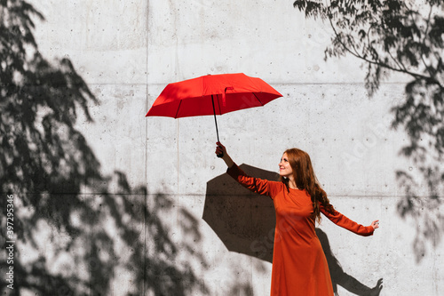 Smiling redhead woman holding umbrella while standing against tree shadow wall photo