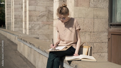 Young woman learning outside near university building, alone young student in sunglasses writing out information from the book. Summer sunny day, preparing for the exam