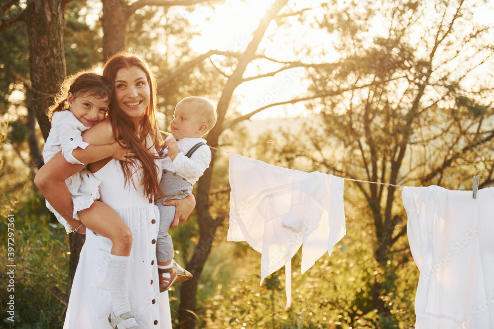 White clothes hanging on the rope to dry. Young mother with her little daughter and son is outdoors in the forest. Beautiful sunshine White clothes hanging on the rope to dry. Young mother with her little daughter and son is outdoors in the forest. Beautiful sunshine