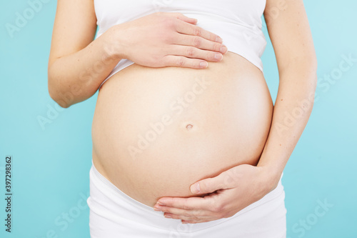 Woman standing and touching with hands her naked big belly. Isolated on blue background. Closeup of pregnant belly.