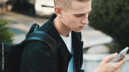 Tracking shot of handsome stylish student guy sitting on bench on street and surfing social networks on smartphone
