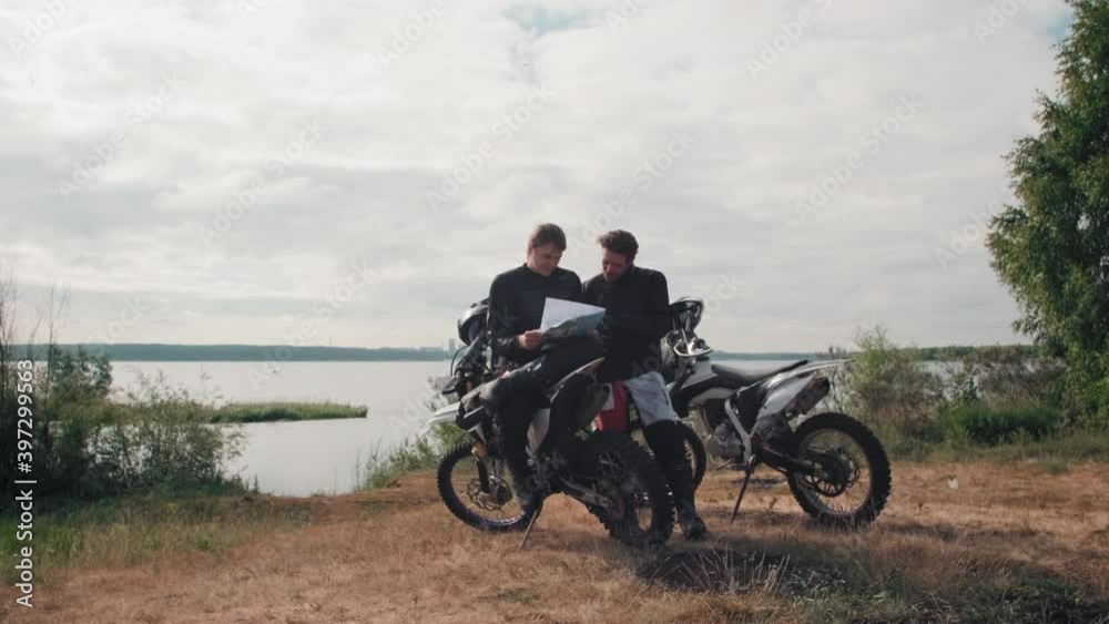 Tracking slowmo of young male motorcyclists resting by lake on gloomy day and looking at map Tracking slowmo of young male motorcyclists resting by lake on gloomy day and looking at map