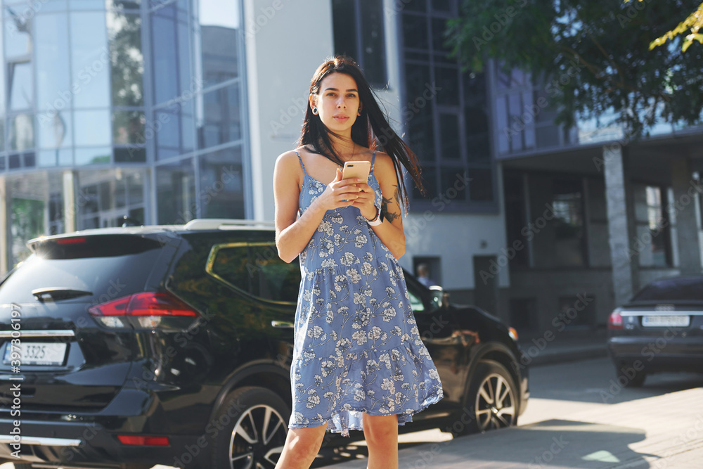Young woman in casual blue dress and with smartphone in hands standing near black car in the city