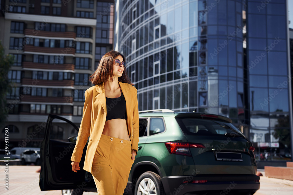 Walks forward from automobile. Young fashionable woman in burgundy colored coat at daytime with her car Walks forward from automobile. Young fashionable woman in burgundy colored coat at daytime with her car