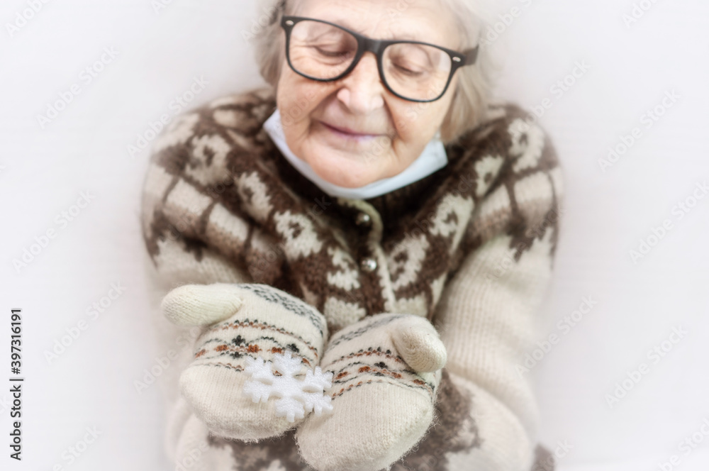 Elderly woman in warm mittens, mask and an Icelandic sweater are holding a large snowflake. Christmas symbols. Winter decor. Celebrating the Christmas holidays. New Year 2021. Coronavirus covid-19 Elderly woman in warm mittens, mask and an Icelandic sweater are holding a large snowflake. Christmas symbols. Winter decor. Celebrating the Christmas holidays. New Year 2021. Coronavirus covid-19