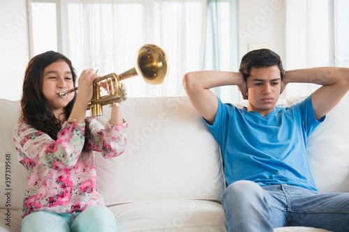 Hispanic brother covering his ears as sister practices trumpet in living room