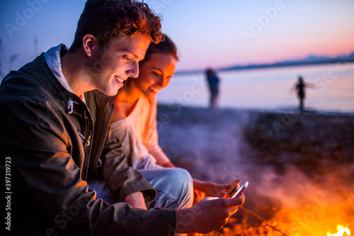 Caucasian couple using cell phone together near fire on beach