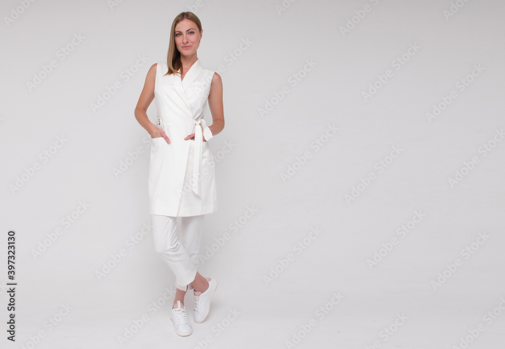 young stylish nurse in white medical costume and white sneakers is standing straight with hands in her pockets and smiling on white wall background. medical concept. free space on right side