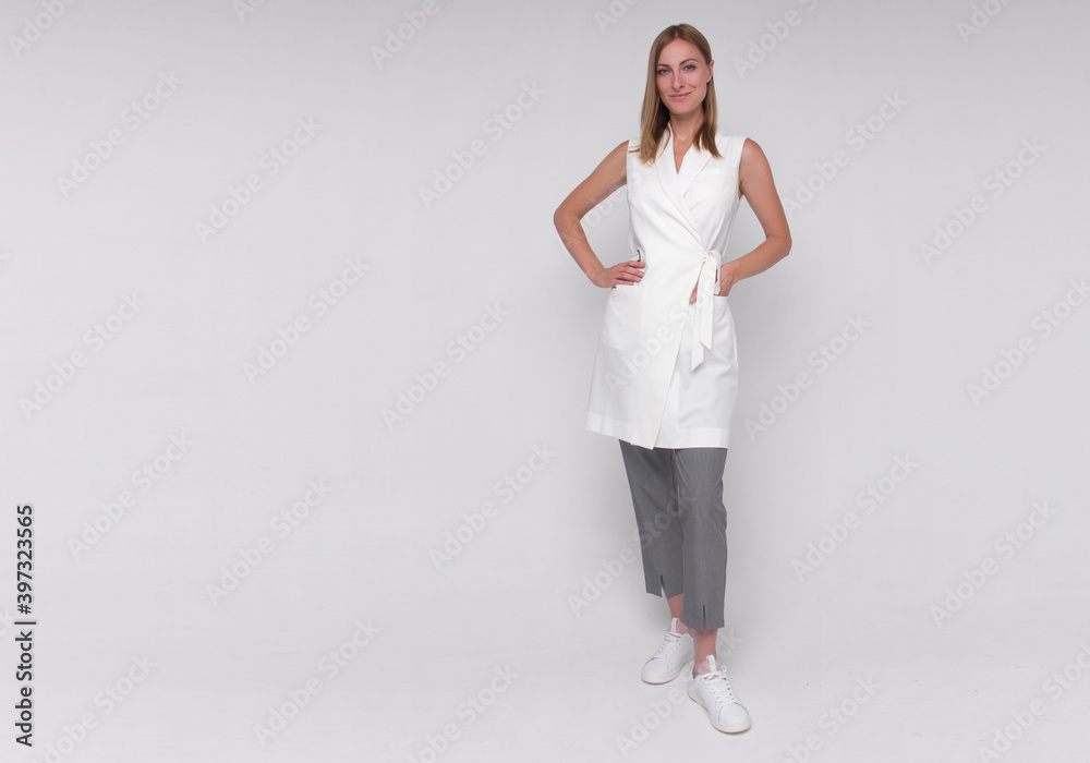 young stylish nurse in white medical costume and gray pants and sneakers is standing straight with hand in her pocket and smiling on white wall background. medical concept. free space on left side young stylish nurse in white medical costume and gray pants and sneakers is standing straight with hand in her pocket and smiling on white wall background. medical concept. free space on left side