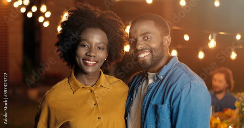 Portrait of young handsome and beautiful African American man and woman smiling cheerfully and standing at outside party at night. Happy guy and girl at evening celebration. Couple in hugs.