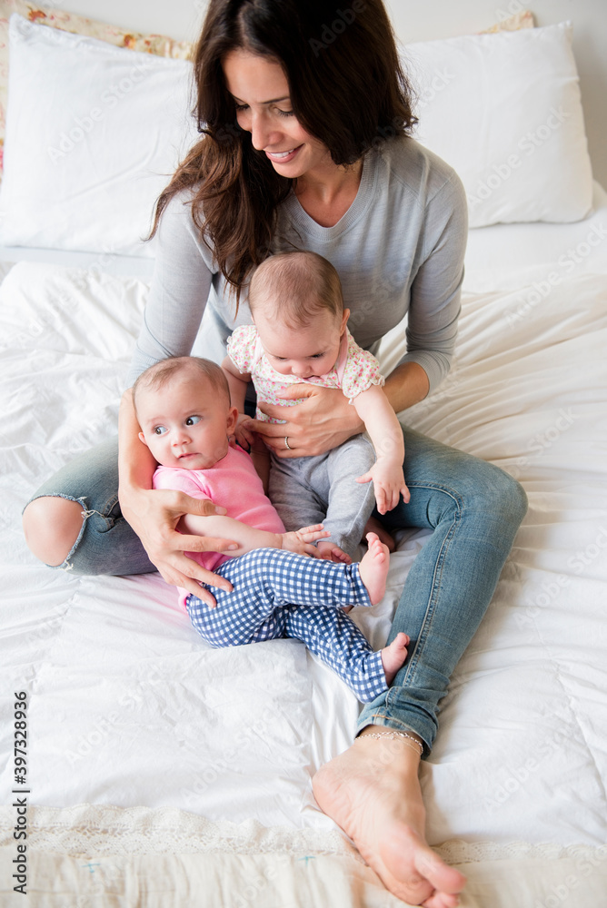 Caucasian mother sitting on bed holding twin baby daughters Caucasian mother sitting on bed holding twin baby daughters