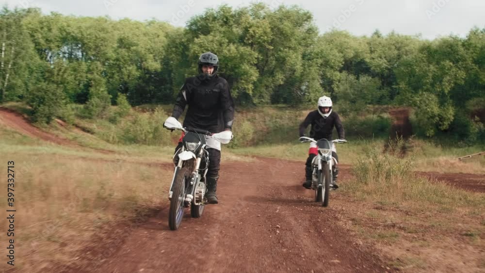 Tracking of two men in helmets and gear riding motorcycles along dirt road in forest Tracking of two men in helmets and gear riding motorcycles along dirt road in forest