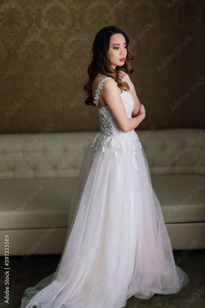 Gorgeous young asian woman in elegant dress looking at camera and window, morning of bride, wedding