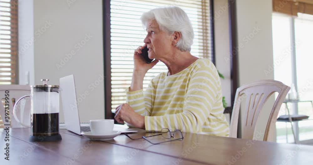 Senior caucasian woman with laptop talking on smartphone at home