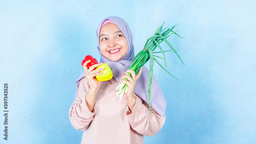 asian hijab woman holds fresh vegetable, eats healthy food at home, uses food product for making vegetarian salad, wears orange jumper, poses indoor. Home growing concept. asian hijab woman holds fresh vegetable, eats healthy food at home, uses food product for making vegetarian salad, wears orange jumper, poses indoor. Home growing concept.