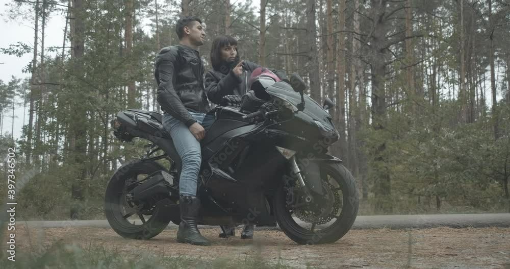 Side view of motorcyclist sitting on black motorbike as smiling woman coming and talking. Wide shot of positive young interracial couple of motorbikers outdoors. Cinema 4k ProRes HQ. Side view of motorcyclist sitting on black motorbike as smiling woman coming and talking. Wide shot of positive young interracial couple of motorbikers outdoors. Cinema 4k ProRes HQ.