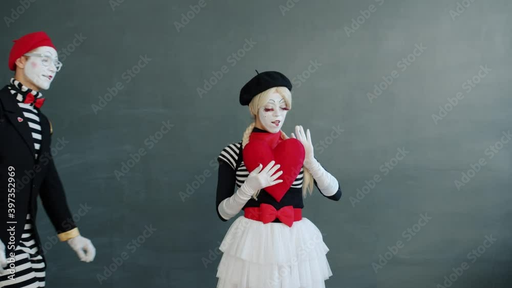Man and woman cute mimes are holding big red heart and kissing standing together on grey background. Romatic relationship and youth concept. Man and woman cute mimes are holding big red heart and kissing standing together on grey background. Romatic relationship and youth concept.