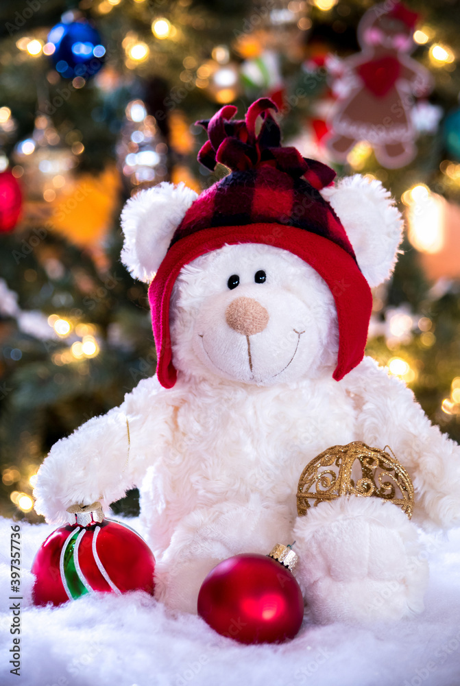 Holiday teddy bear with red hat, holds Christmas ornaments in front of a lit Christmas tree 