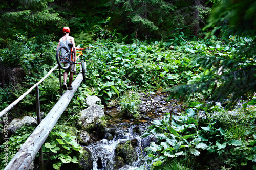 Mountain biker holding a bicycle in his hands crossing a mountain river along a log.