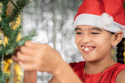 happy smiling girl kid in santa hat decorating Christmas tree for festive holyday celebration at home.
