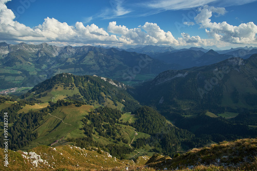 Aerial panoramic view of beautiful countryside with green hills and mountain ranges in the background and cloudy blue sky above on a sunny day in Chur, Switzerland. © Matthew