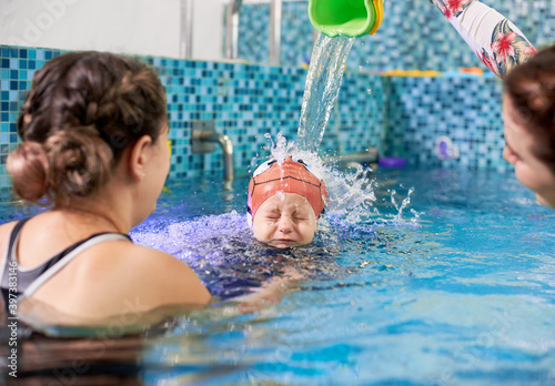 Woman holding boy while female instructor pouring water on kid head. Loving mother spending time with lovely child in swimming pool with crystal blue water. Concept of safe swimming training.