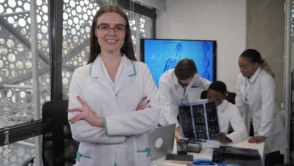 Beautiful female doctor in glasses folds her arms and smiles at the camera on the background of multinational doctors in a modern hospital. Healthcare, medicine concept. Beautiful female doctor in glasses folds her arms and smiles at the camera on the background of multinational doctors in a modern hospital. Healthcare, medicine concept.
