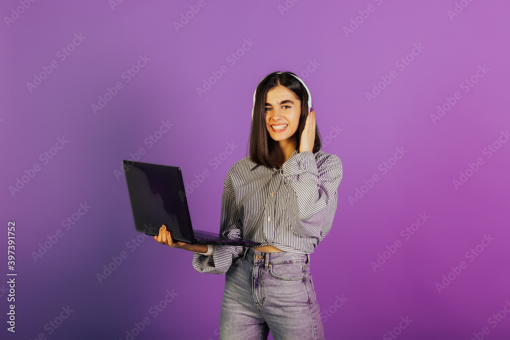 Happy girl in headphones smiling while holding laptop isolated over violet background.
