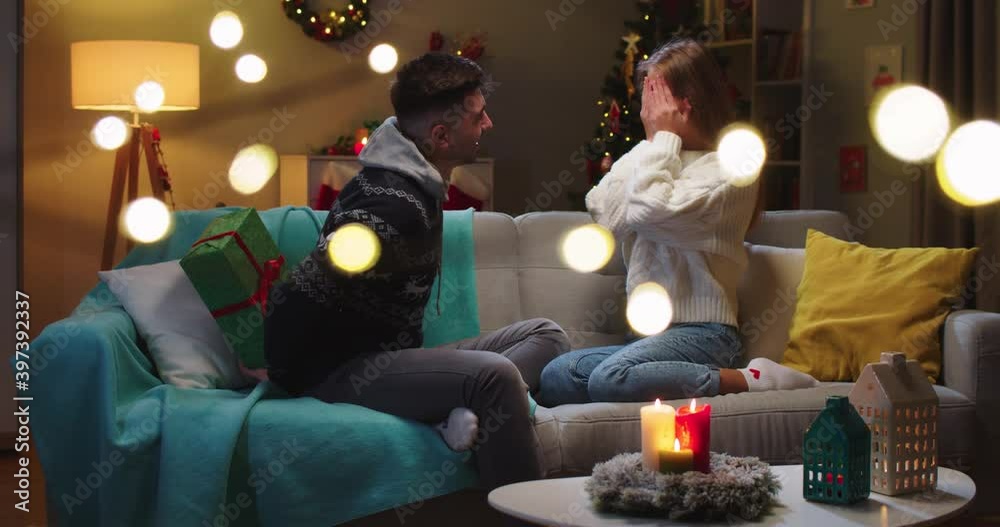 Middle plan of happy couple husband and wife sitting on sofa. Girlfriend sitting with closed eyes waiting for present. Boyfriend giving big beautiful Christmas present. Concept of family holidays. Middle plan of happy couple husband and wife sitting on sofa. Girlfriend sitting with closed eyes waiting for present. Boyfriend giving big beautiful Christmas present. Concept of family holidays.