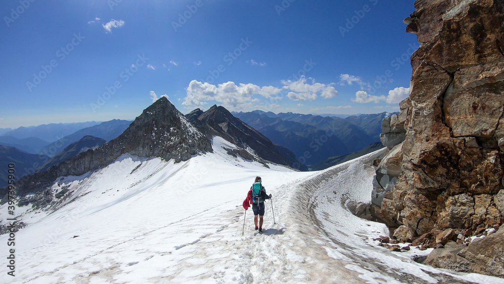 A woman with hiking stick hiking through a glacier in Heiligenblut region in Austria, on the way to Hohe Sonnblick. There are many high chains of Alps behind her. Sunny day. Adventure and discovery.