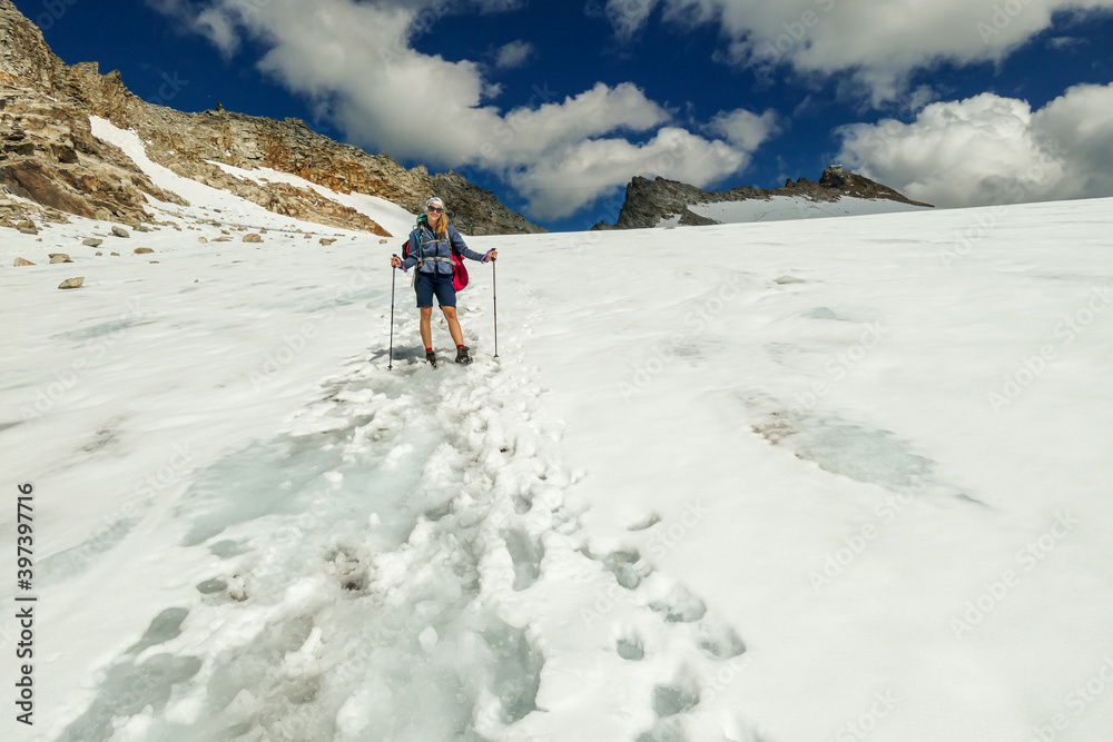 A woman following the narrow footprints path while crossing the glacier under Hohe Sonnblick in Austrian Alps. The footprints lead in one direction. Many high mountain chains in the nearby. Sunny day