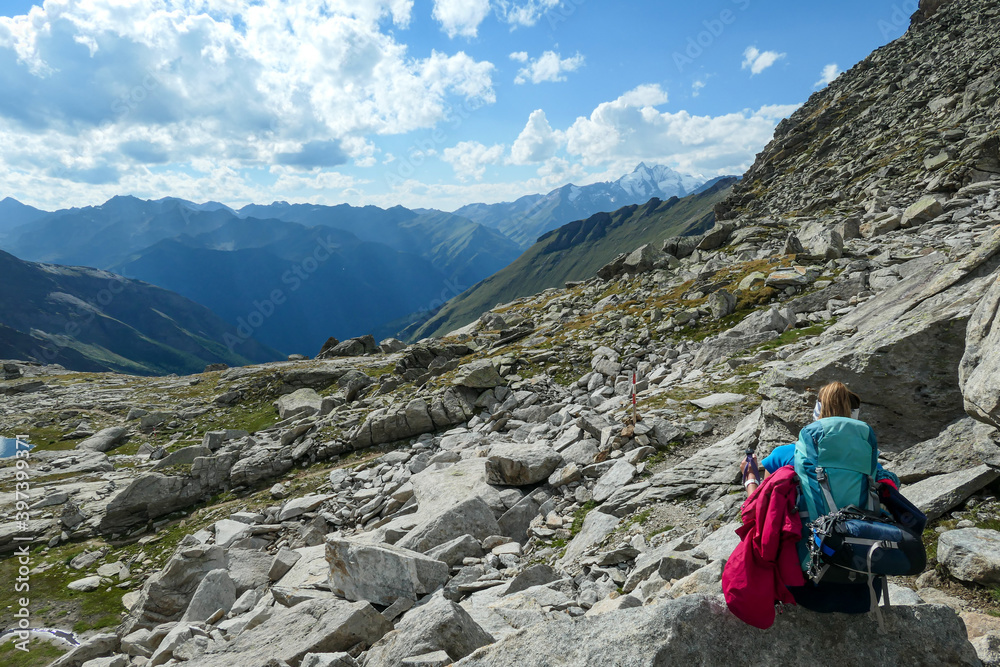 Fototapeta premium Panoramic view from Hohe Sonnblick in Austrian Alps on Gro?glockner. A woman with hiking backpack resting next to a narrow pathway through the stony landscape, full of lose stones and boulders.