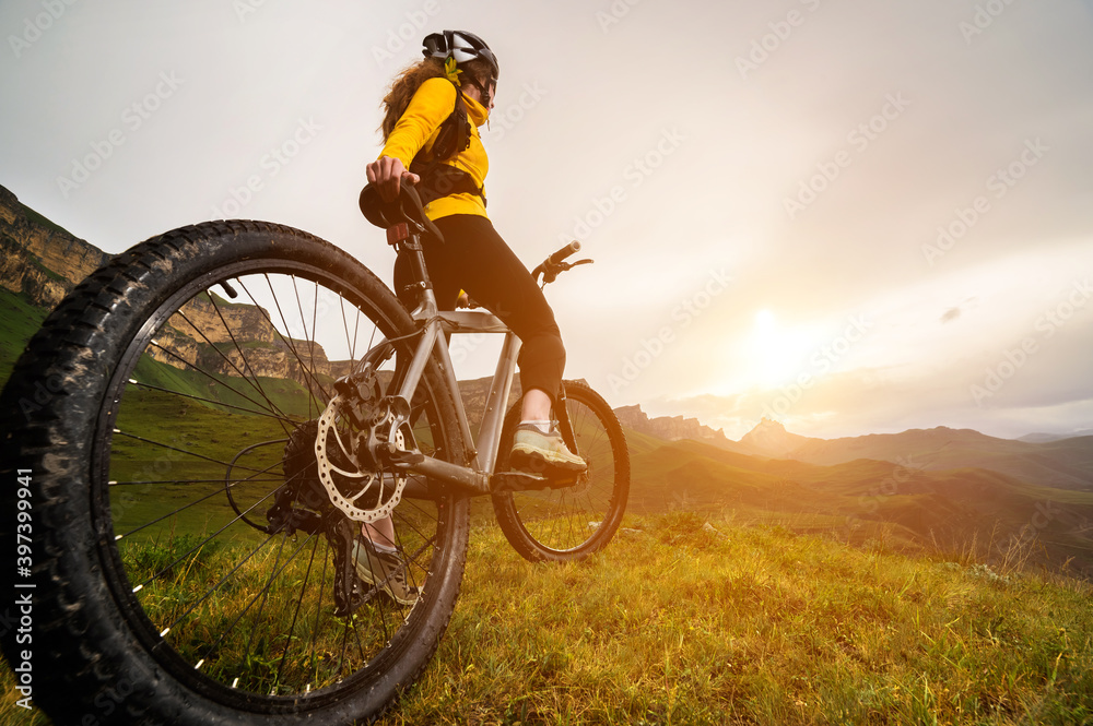 Obraz premium Wide angle bottom view of a young woman sitting on a mountain bike high in the mountains against the backdrop of epic rocks in the evening. Mountain sports bike