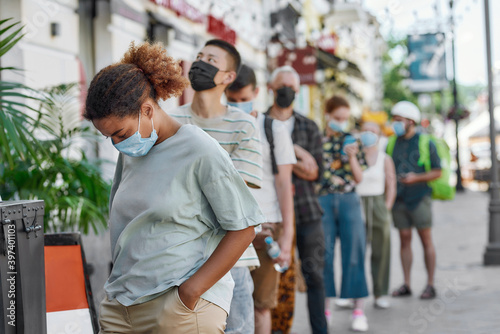Young african american woman wearing mask waiting, standing in line with other people, respecting social distancing to collect takeaway order from the pickup point during lockdown