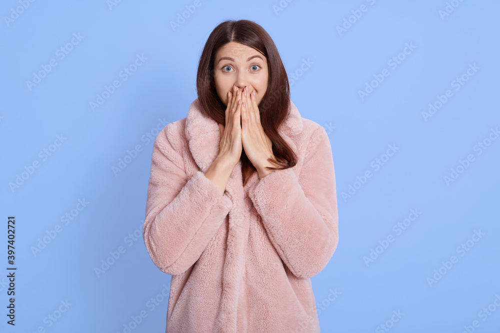Winsome young female has overjoyed expression, covers mouth with both palms, has eyes popped out, dressed in stylish pale pink fur coat, stands against blue background. Winsome young female has overjoyed expression, covers mouth with both palms, has eyes popped out, dressed in stylish pale pink fur coat, stands against blue background.